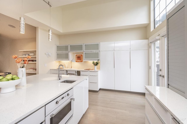 kitchen with light stone counters, oven, decorative light fixtures, white cabinetry, and a sink