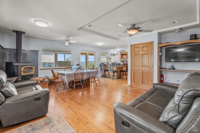 living room with ceiling fan, a wood stove, light hardwood / wood-style flooring, and a raised ceiling