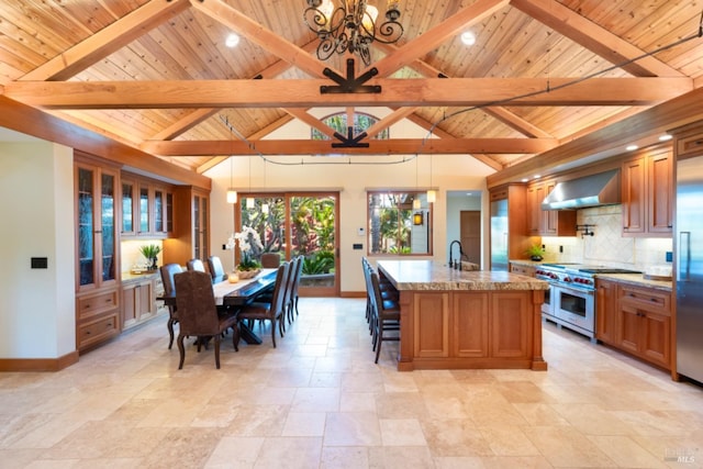 kitchen with hanging light fixtures, light stone counters, premium appliances, an island with sink, and wall chimney exhaust hood