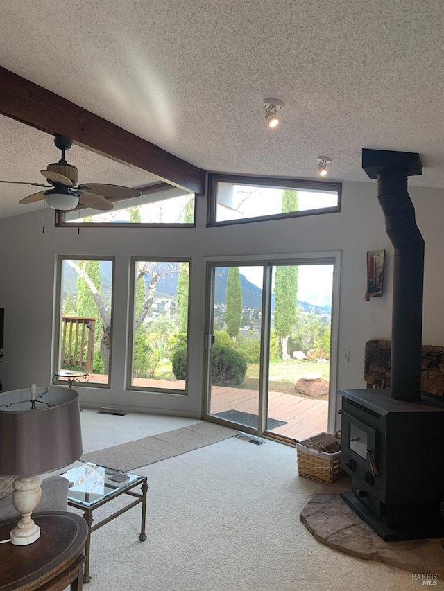 carpeted living room with ceiling fan, a wood stove, vaulted ceiling with beams, and a textured ceiling