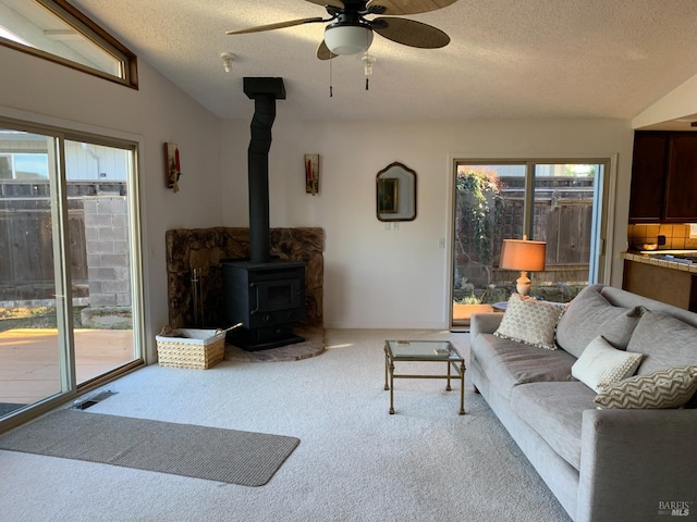 carpeted living room with ceiling fan, vaulted ceiling, a textured ceiling, and a wood stove