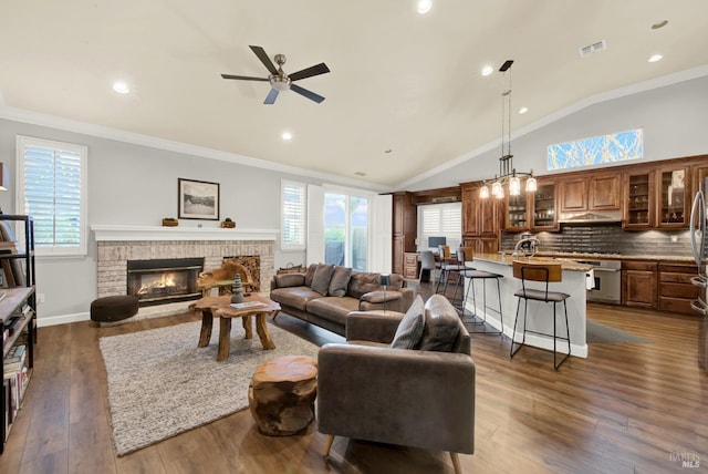 living room featuring dark wood-type flooring, lofted ceiling, a brick fireplace, ornamental molding, and ceiling fan