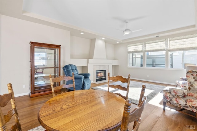 dining room featuring a tiled fireplace, light hardwood / wood-style flooring, a raised ceiling, and ceiling fan