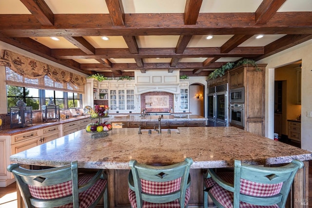 kitchen featuring a breakfast bar area, a large island with sink, coffered ceiling, built in appliances, and light stone counters