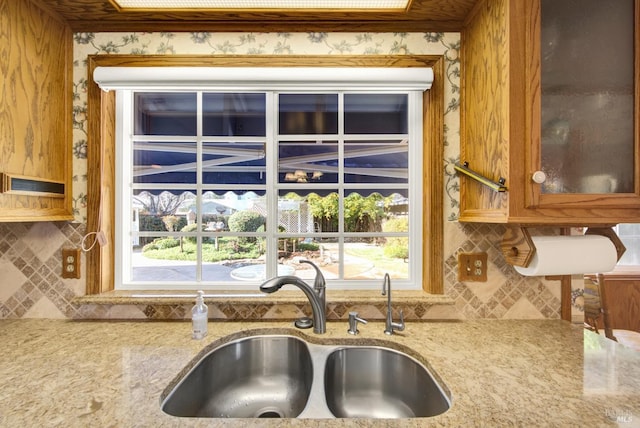 details featuring tasteful backsplash, light stone counters, a sink, and glass insert cabinets