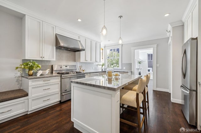 kitchen featuring wall chimney range hood, appliances with stainless steel finishes, white cabinetry, a kitchen island, and decorative light fixtures