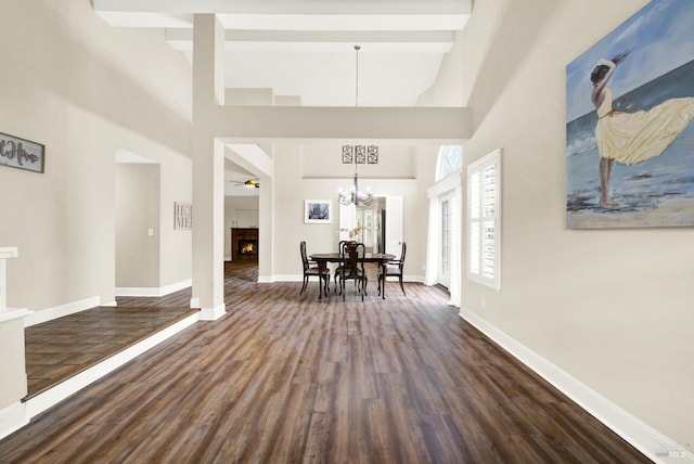 unfurnished dining area featuring dark wood-type flooring, a towering ceiling, beam ceiling, and ceiling fan with notable chandelier