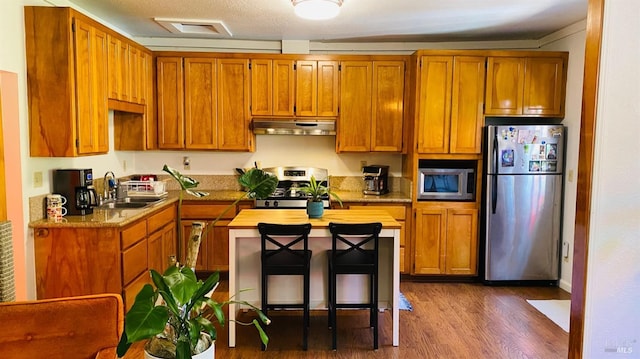 kitchen with appliances with stainless steel finishes, a breakfast bar, sink, light stone counters, and dark wood-type flooring