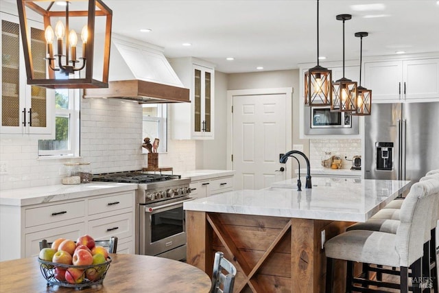 kitchen featuring high end appliances, premium range hood, a center island with sink, and white cabinets