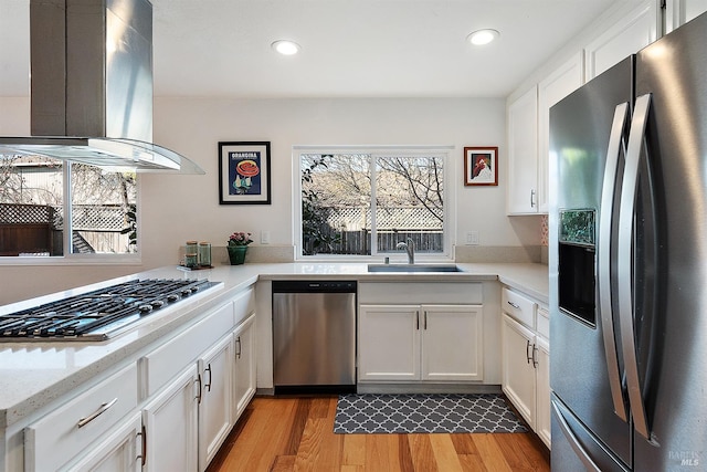 kitchen with appliances with stainless steel finishes, island range hood, white cabinetry, sink, and light hardwood / wood-style floors