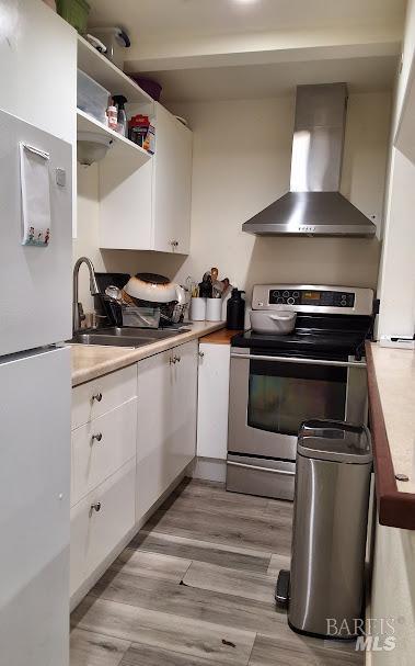 kitchen with wall chimney exhaust hood, sink, stainless steel range with electric cooktop, refrigerator, and white cabinets