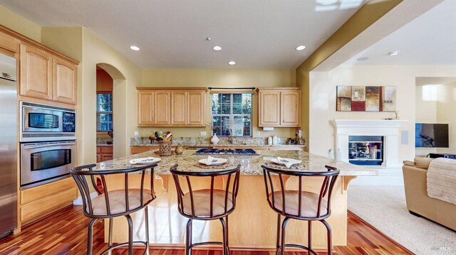 kitchen with light brown cabinetry, light stone counters, appliances with stainless steel finishes, a kitchen island, and hardwood / wood-style floors