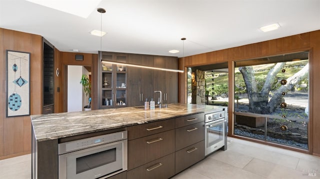 kitchen featuring sink, a kitchen island with sink, hanging light fixtures, stainless steel appliances, and wood walls