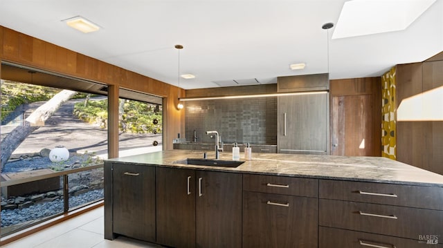 kitchen with sink, dark brown cabinets, light stone counters, decorative light fixtures, and wood walls