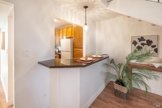 kitchen with dark wood-type flooring, dark stone countertops, kitchen peninsula, white fridge, and pendant lighting