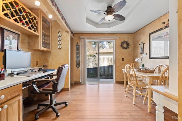 home office with ceiling fan, light wood-type flooring, and a tray ceiling