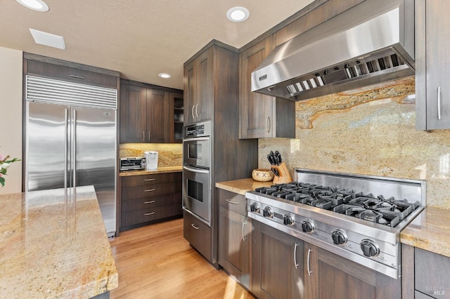 kitchen with stainless steel appliances, light stone counters, light hardwood / wood-style floors, decorative backsplash, and wall chimney exhaust hood