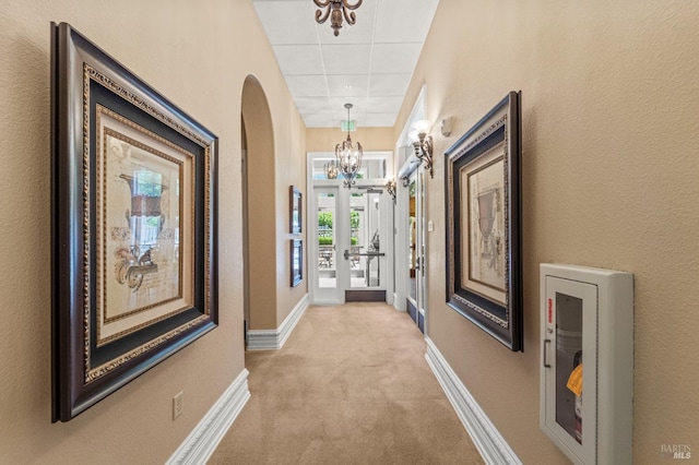 hall featuring arched walkways, a drop ceiling, light colored carpet, baseboards, and an inviting chandelier