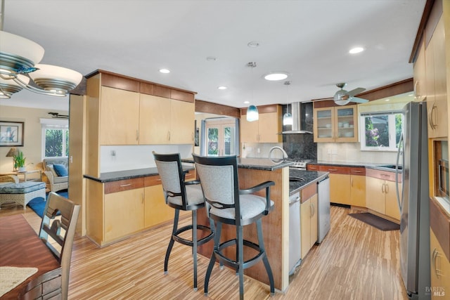 kitchen featuring wall chimney exhaust hood, a center island with sink, light brown cabinets, appliances with stainless steel finishes, and light hardwood / wood-style floors