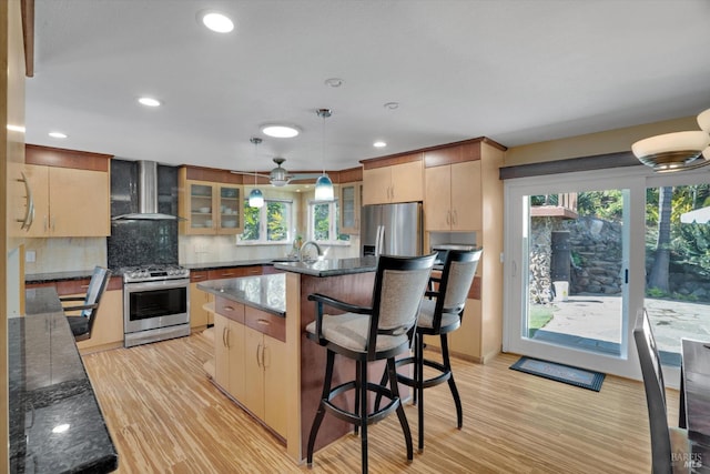 kitchen featuring wall chimney exhaust hood, stainless steel appliances, decorative light fixtures, and light brown cabinets