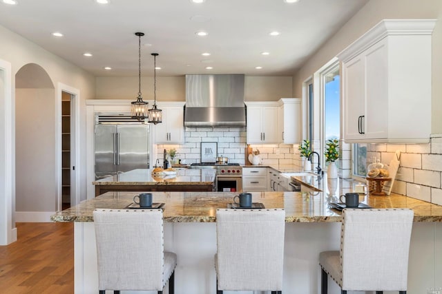 kitchen featuring high end appliances, light stone counters, a center island, wall chimney range hood, and white cabinets