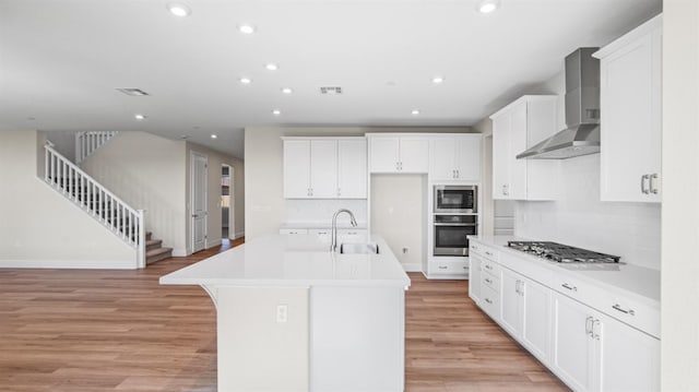 kitchen with stainless steel appliances, light countertops, a kitchen island with sink, a sink, and wall chimney exhaust hood