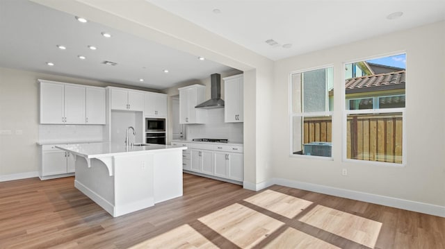 kitchen with oven, an island with sink, wall chimney exhaust hood, and white cabinetry