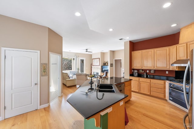 kitchen featuring oven, a kitchen island with sink, light brown cabinetry, a sink, and dark countertops