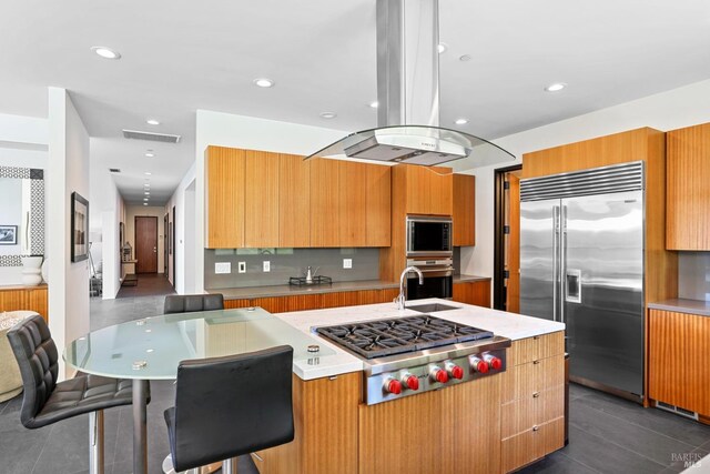 kitchen with island range hood, built in appliances, an island with sink, and dark tile patterned floors