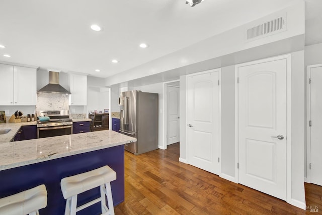 kitchen featuring wall chimney range hood, stainless steel appliances, light stone counters, white cabinets, and a kitchen bar