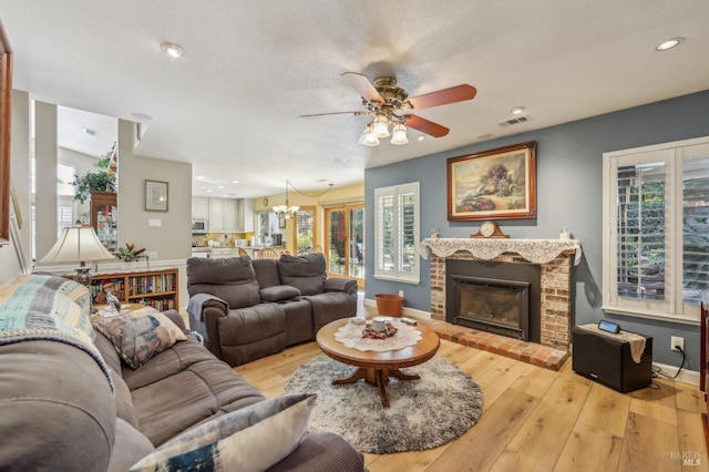 living room with ceiling fan with notable chandelier, a textured ceiling, a fireplace, and light hardwood / wood-style floors