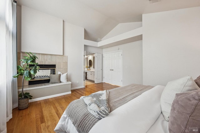 bedroom with wood finished floors, baseboards, ensuite bath, a tile fireplace, and vaulted ceiling