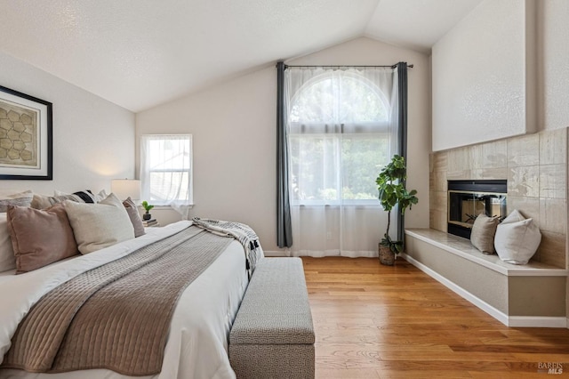 bedroom with a tiled fireplace, light wood finished floors, baseboards, and vaulted ceiling