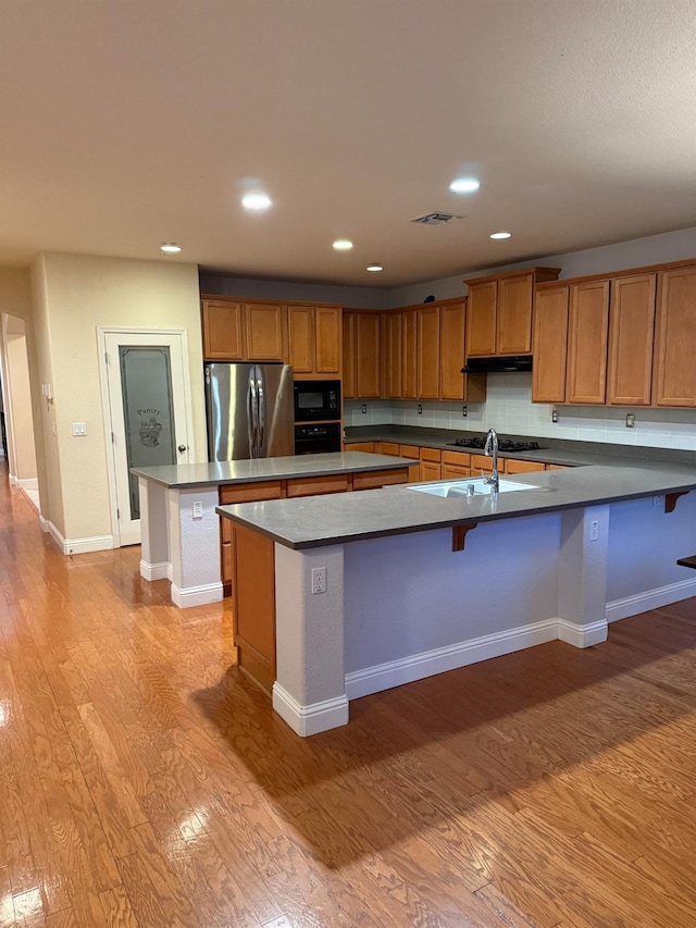 kitchen featuring under cabinet range hood, a peninsula, black appliances, light wood finished floors, and brown cabinetry