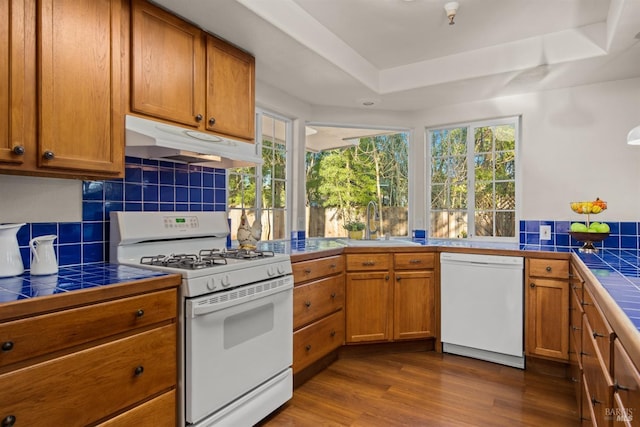 kitchen featuring tile countertops, brown cabinetry, a sink, white appliances, and under cabinet range hood