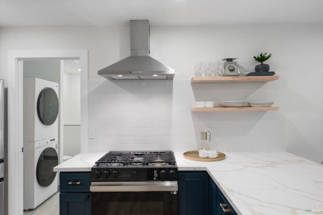 kitchen with stacked washer and dryer, blue cabinets, tasteful backsplash, gas range oven, and wall chimney range hood