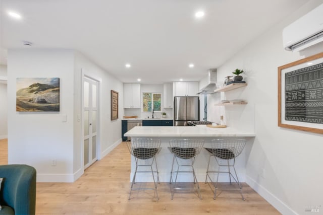 kitchen with appliances with stainless steel finishes, kitchen peninsula, a wall unit AC, white cabinets, and wall chimney range hood