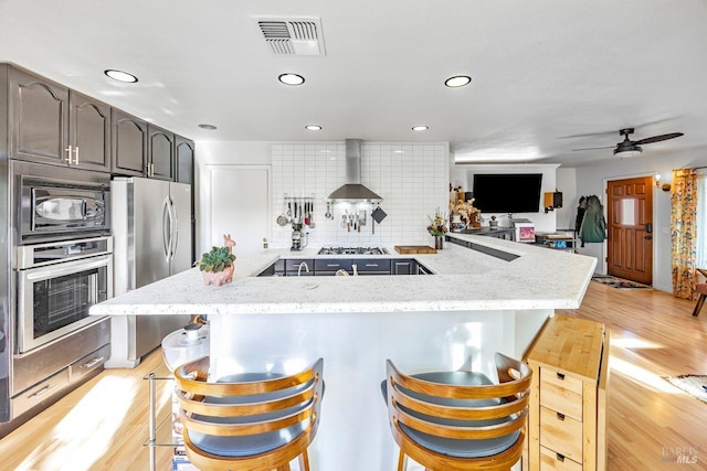 kitchen featuring a breakfast bar area, appliances with stainless steel finishes, light stone countertops, kitchen peninsula, and wall chimney exhaust hood