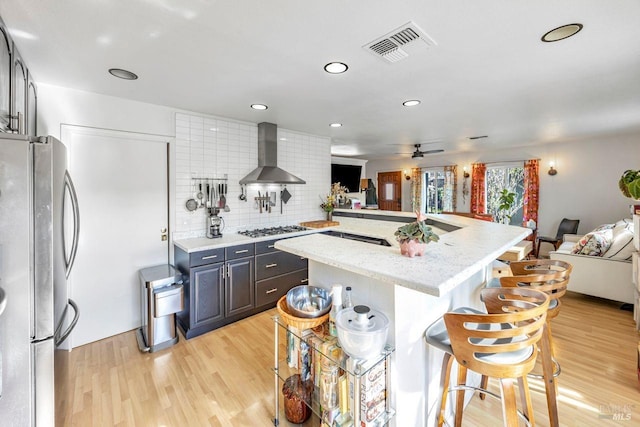 kitchen with a kitchen bar, stainless steel appliances, light stone countertops, light wood-type flooring, and wall chimney exhaust hood