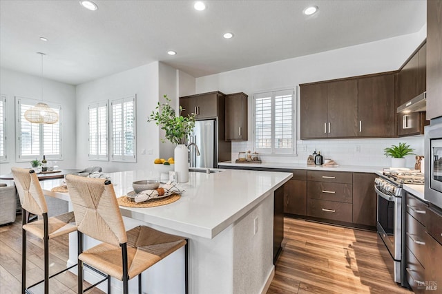 kitchen with a center island with sink, stainless steel appliances, light countertops, dark brown cabinets, and light wood-type flooring