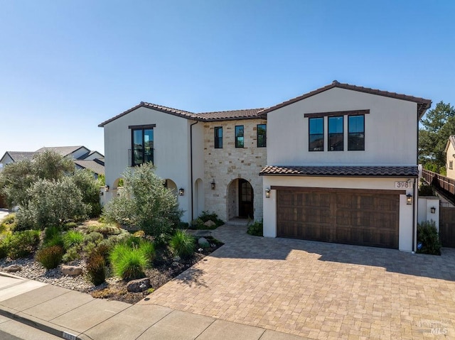 mediterranean / spanish-style house with decorative driveway, a tile roof, stucco siding, an attached garage, and stone siding