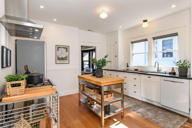 kitchen featuring light wood-style flooring, a sink, wall chimney range hood, dishwasher, and dark countertops