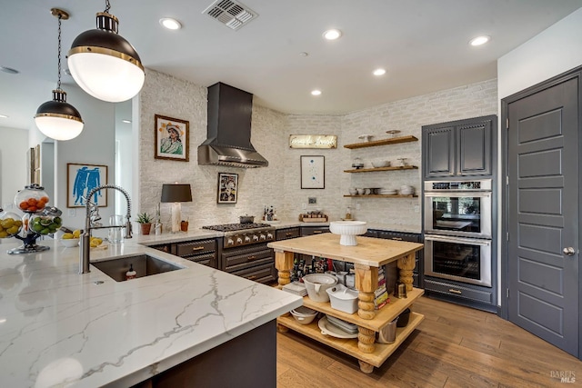 kitchen with wall chimney exhaust hood, hanging light fixtures, light stone countertops, open shelves, and a sink