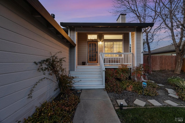 exterior entry at dusk featuring fence, a chimney, and covered porch