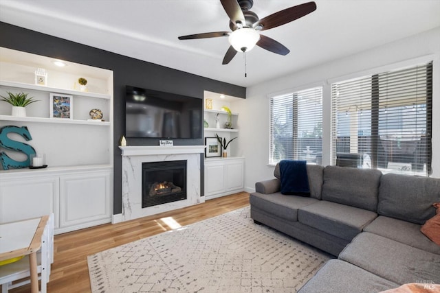 living room with built in features, a premium fireplace, ceiling fan, and light wood-type flooring