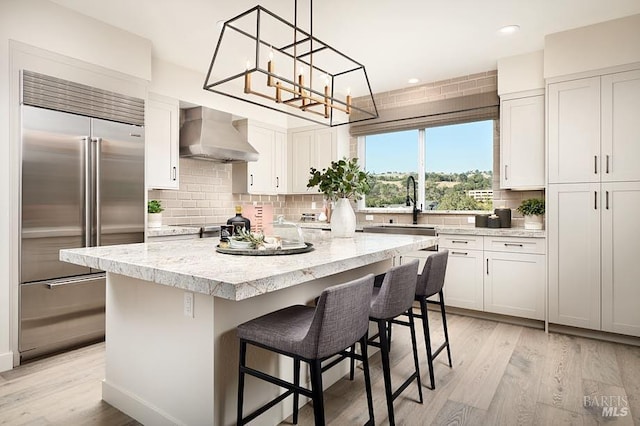 kitchen featuring wall chimney range hood, a kitchen island, white cabinets, and built in refrigerator