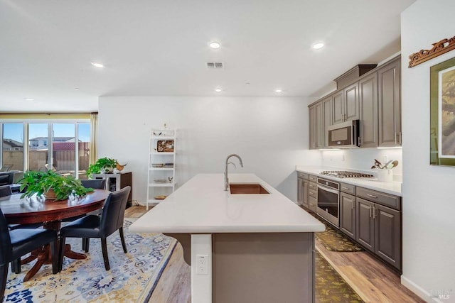 kitchen with sink, a kitchen island with sink, stainless steel appliances, a kitchen bar, and light wood-type flooring