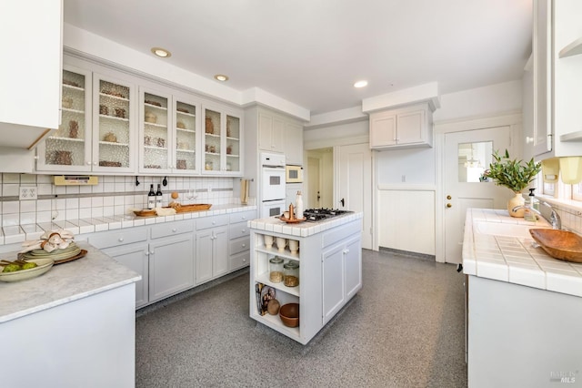 kitchen with tile counters, a sink, tasteful backsplash, open shelves, and glass insert cabinets