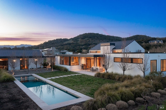 back house at dusk featuring a fenced in pool, a mountain view, a lawn, and a patio area