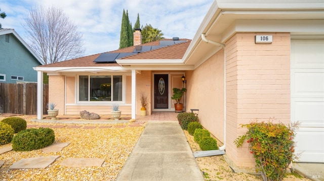 entrance to property featuring a porch, a shingled roof, fence, roof mounted solar panels, and a chimney